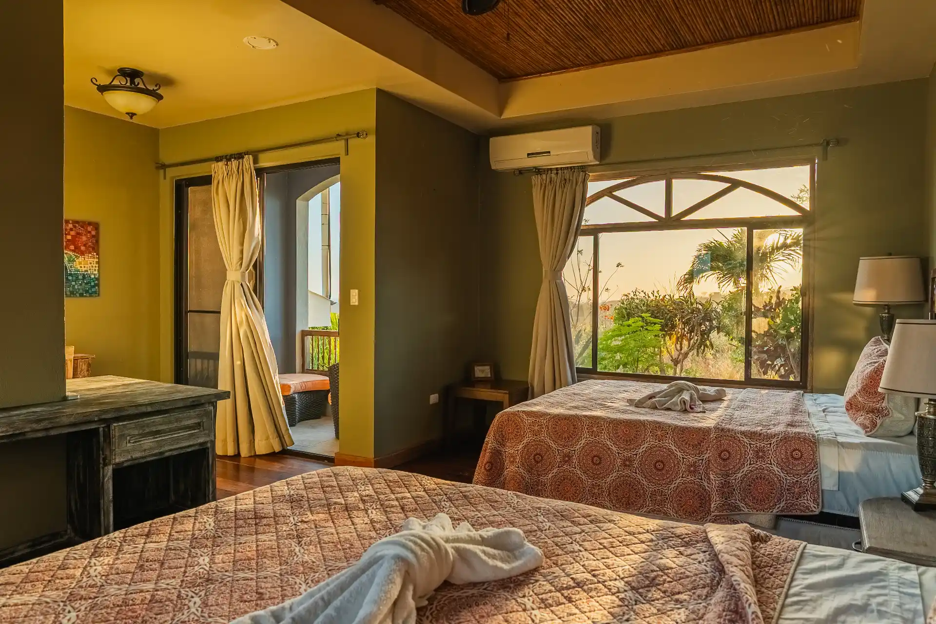 Cozy bedroom with two beds, patterned bedspreads, air conditioning, curtained windows, and tropical greenery visible outside.