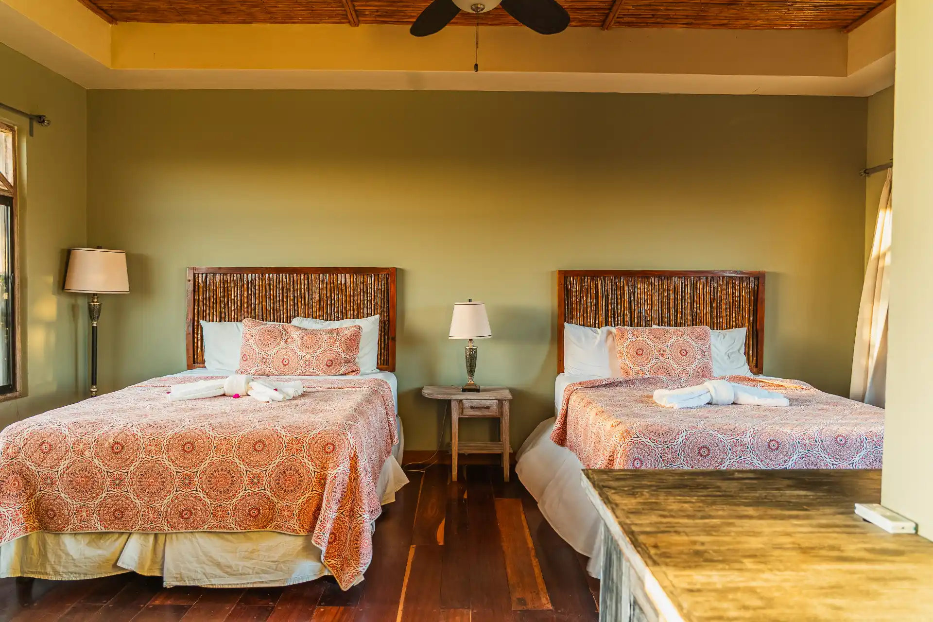 Bedroom with two beds, patterned bedspreads, woven wood headboards, bedside table with lamp, and wooden flooring.