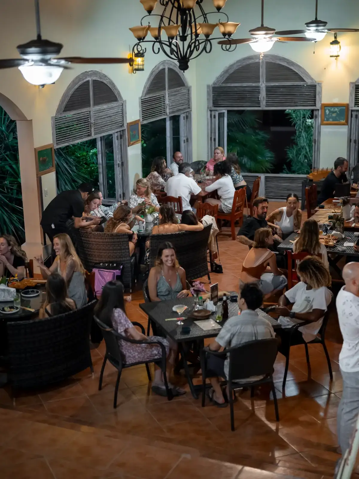 Group of people dining and socializing together at tables inside a restaurant with large open windows and tropical greenery outside