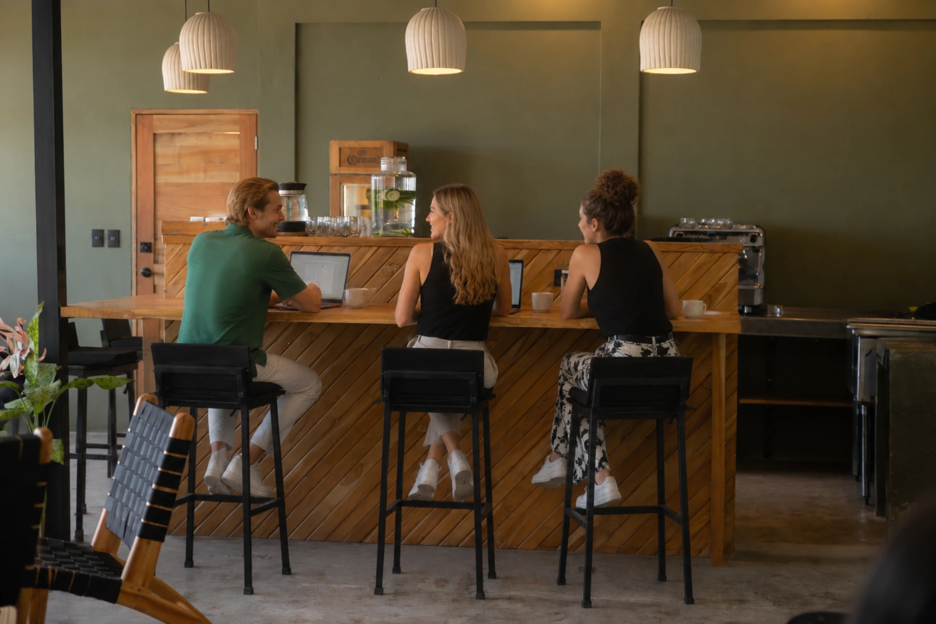 Three people working on laptops at a wooden counter with stools, pendant lights, and a coffee station.