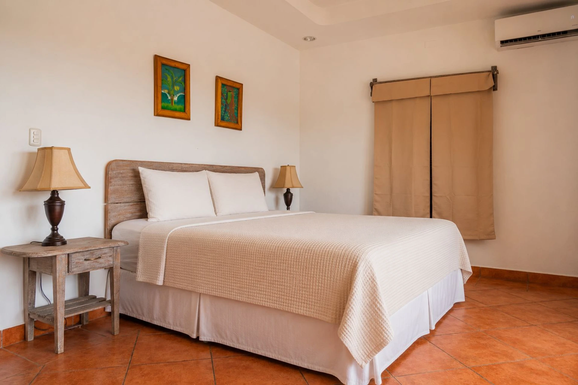 King Standard Room with a bed, wooden bedside tables and lamps, framed artwork, beige curtains, and terracotta tile flooring