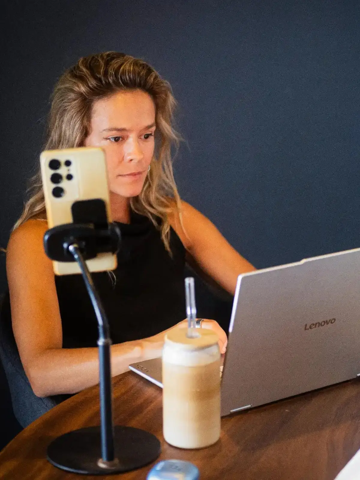 Woman working on a laptop at a wooden table with a smartphone mounted on a stand and a drink beside her